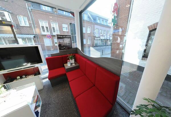 Heller Wartebereich mit roten Sitzbänken, großem Fenster und Blick auf eine Apotheke.